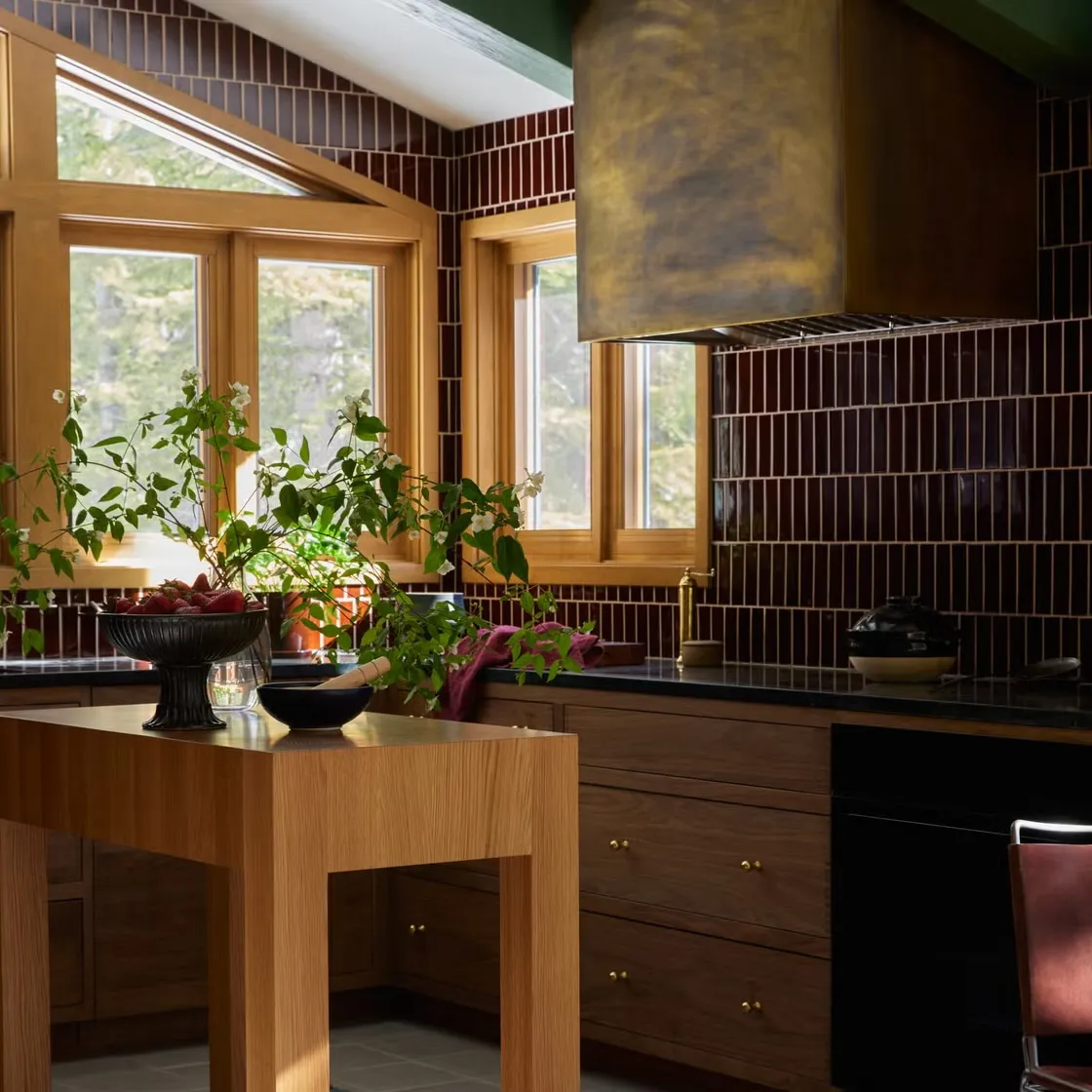 Kitchen corner with oxblood tiled walls, a brass range hood and walnut drawers, an oak island holding strawberries and a flowering branch.
