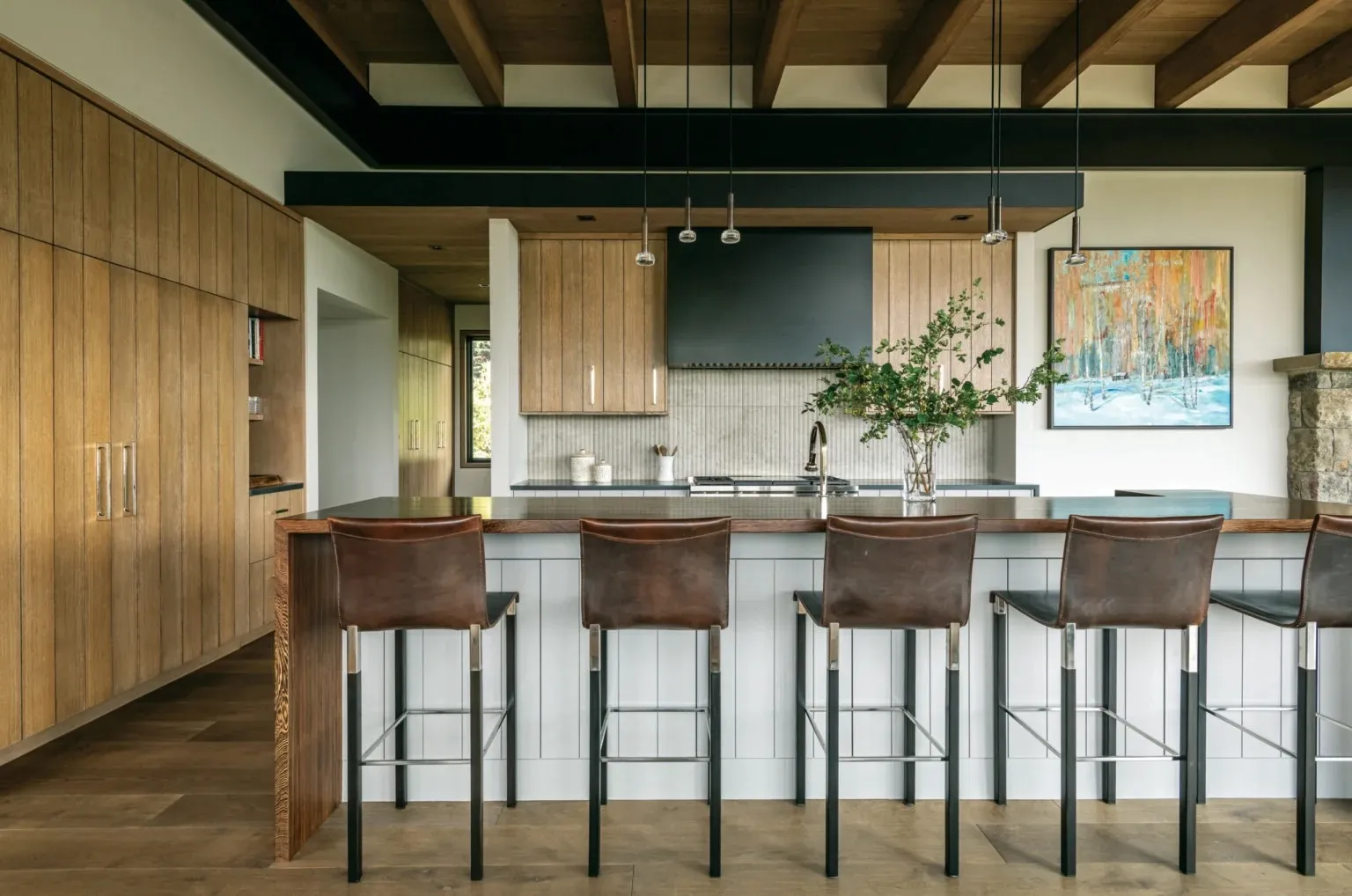 Kitchen with oak cabinetry, a long walnut-topped island lined with leather bar stools, and a black range hood under exposed timber beams.