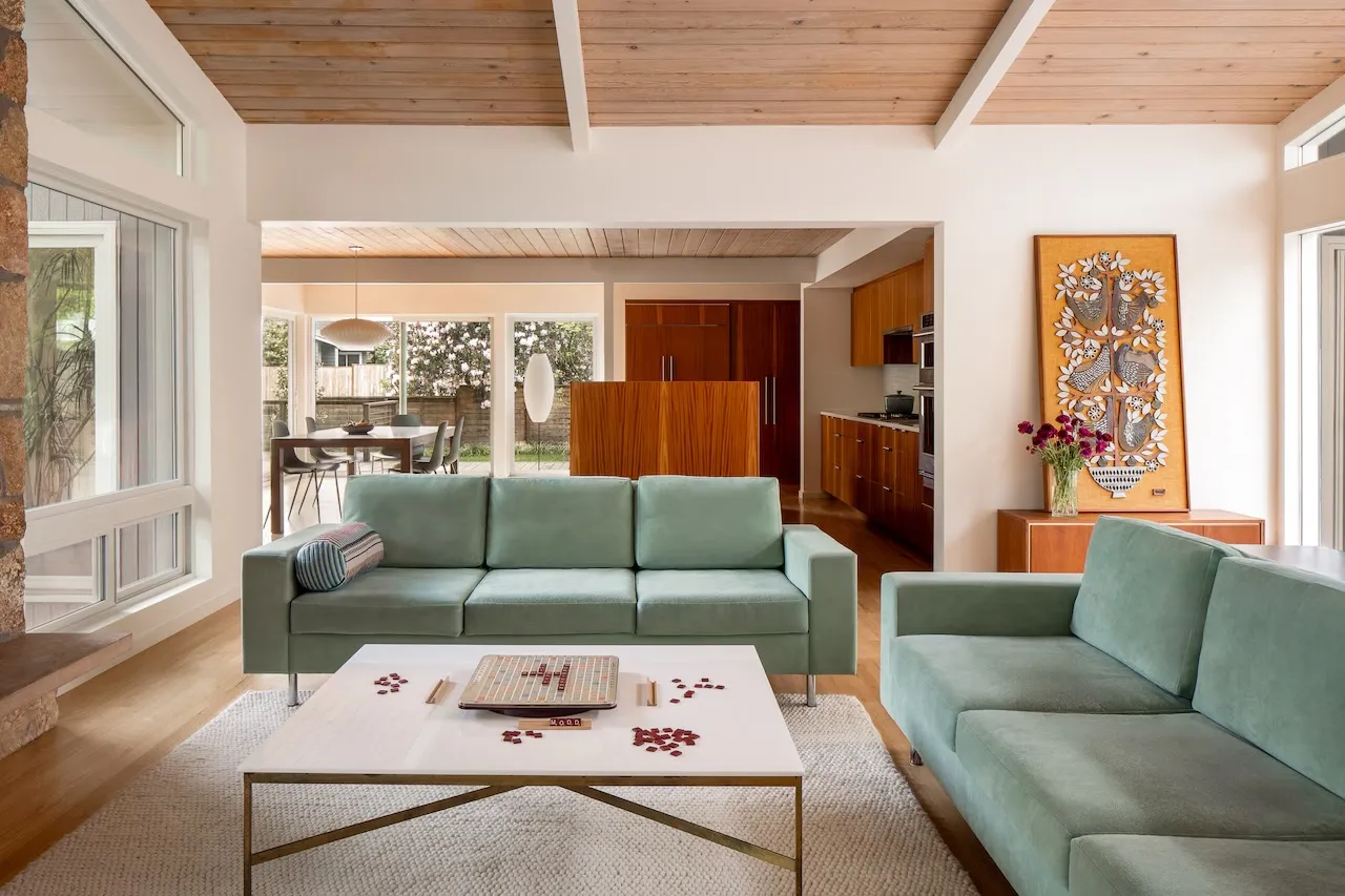 Living room with two sage-green sofas under a pale timber ceiling, opening through to the dining area and a teak kitchen.