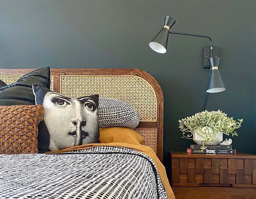 Bed with a cane-and-walnut headboard and patterned cushions against a dark green wall, a black swing-arm lamp above the nightstand.