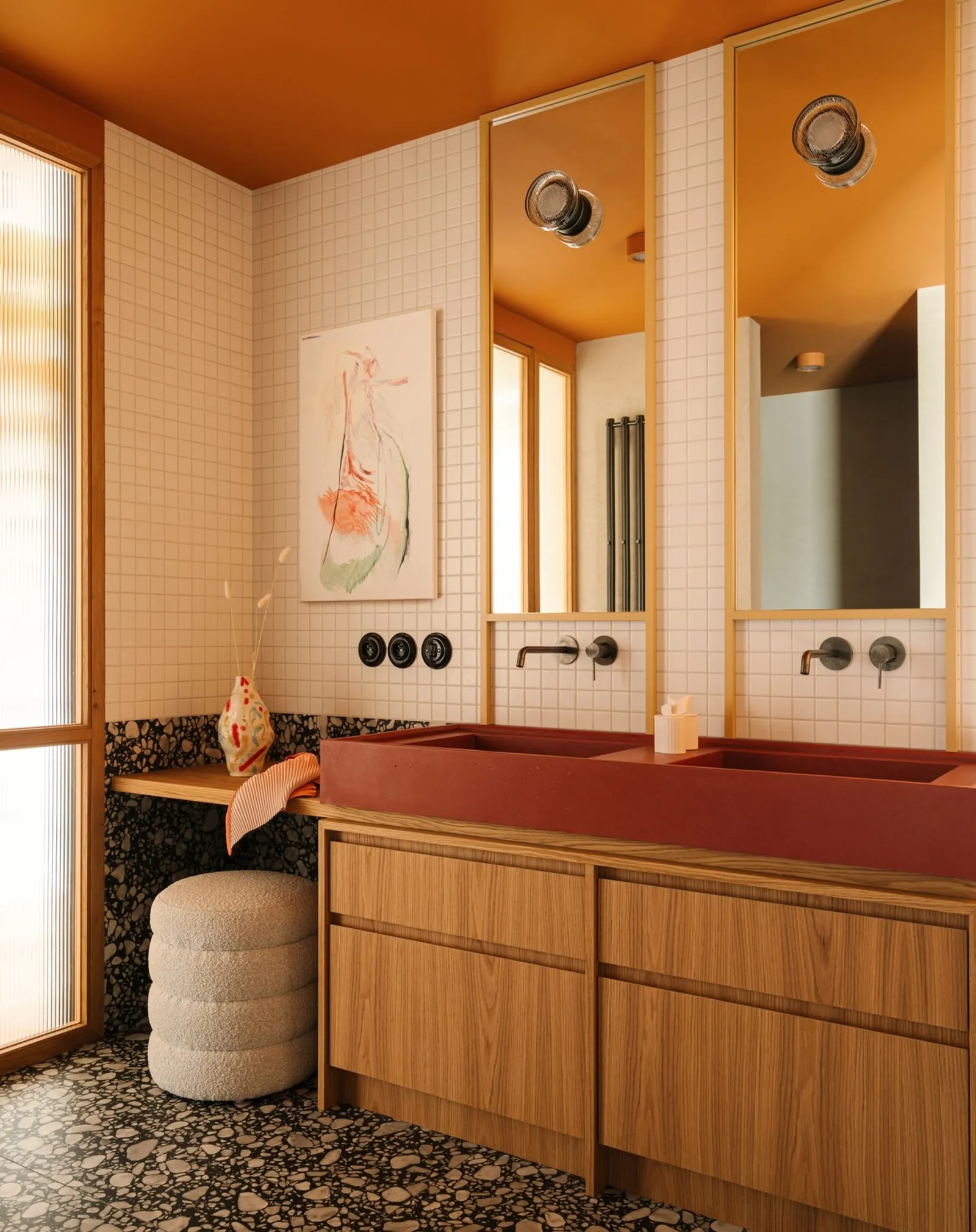 Bathroom with an oxblood double trough sink on an oak vanity, tall brass-framed mirrors and terrazzo floor under an ochre ceiling.