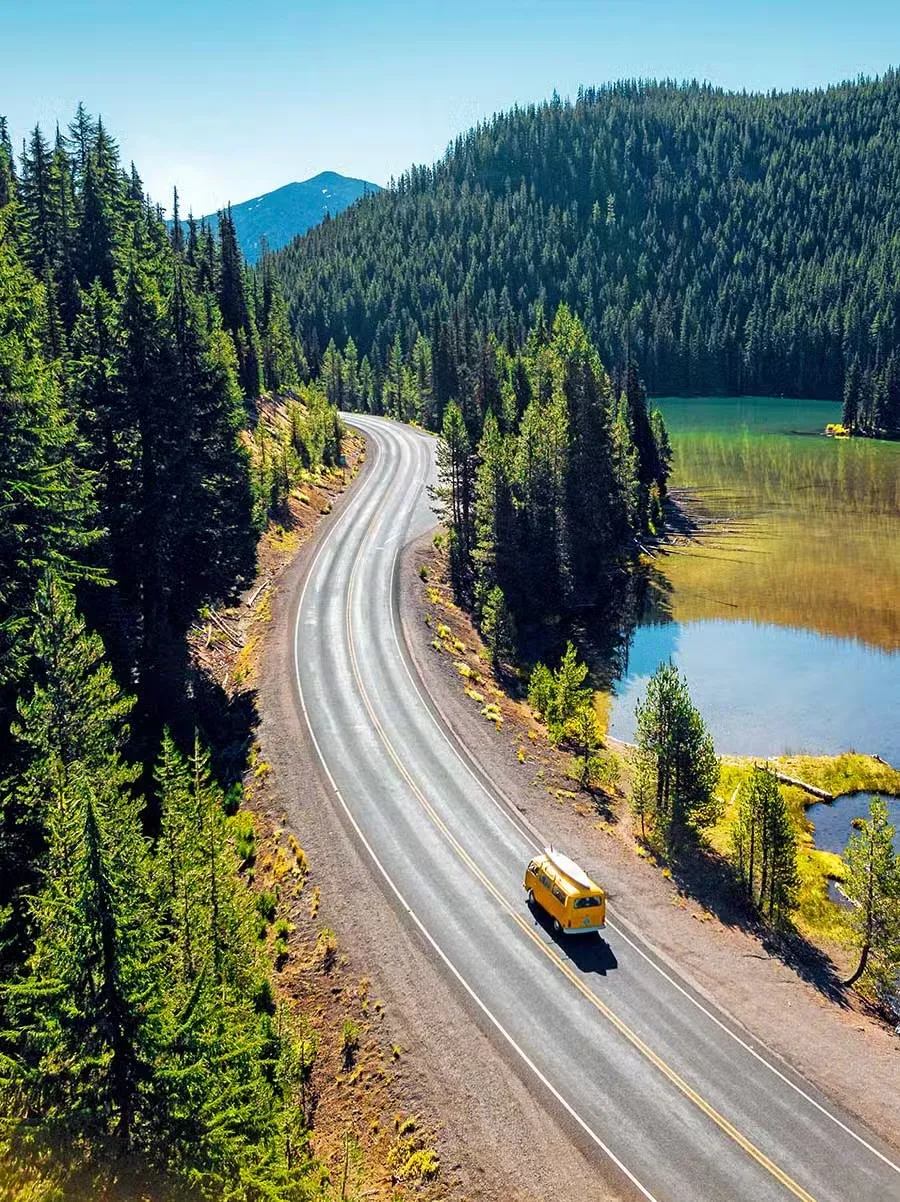 Aerial view of a yellow camper van on a winding road through conifer forest, past a still green lake.
