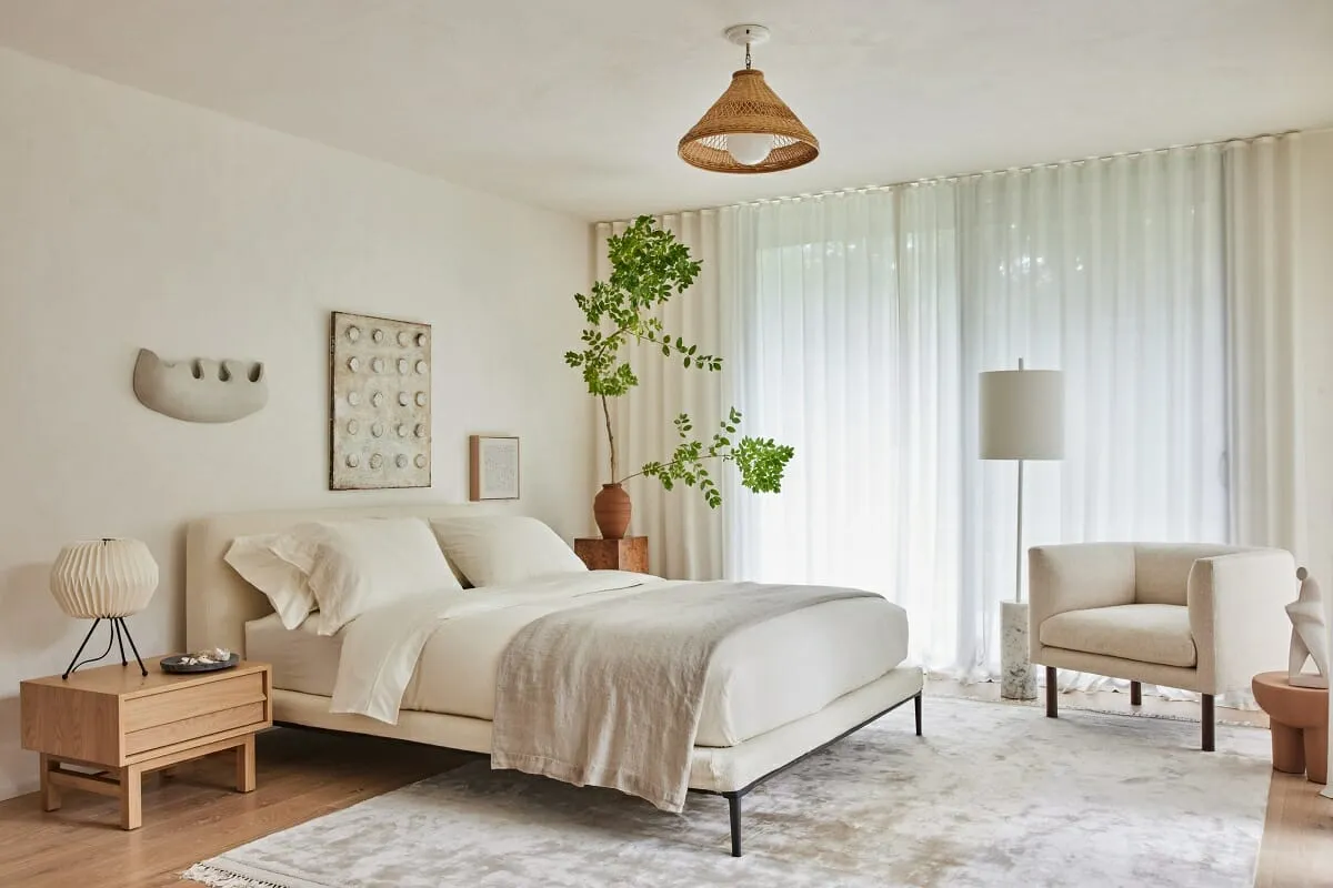 Pale bedroom with a low upholstered bed, a woven pendant and a branching tree in a clay pot before sheer full-height curtains.
