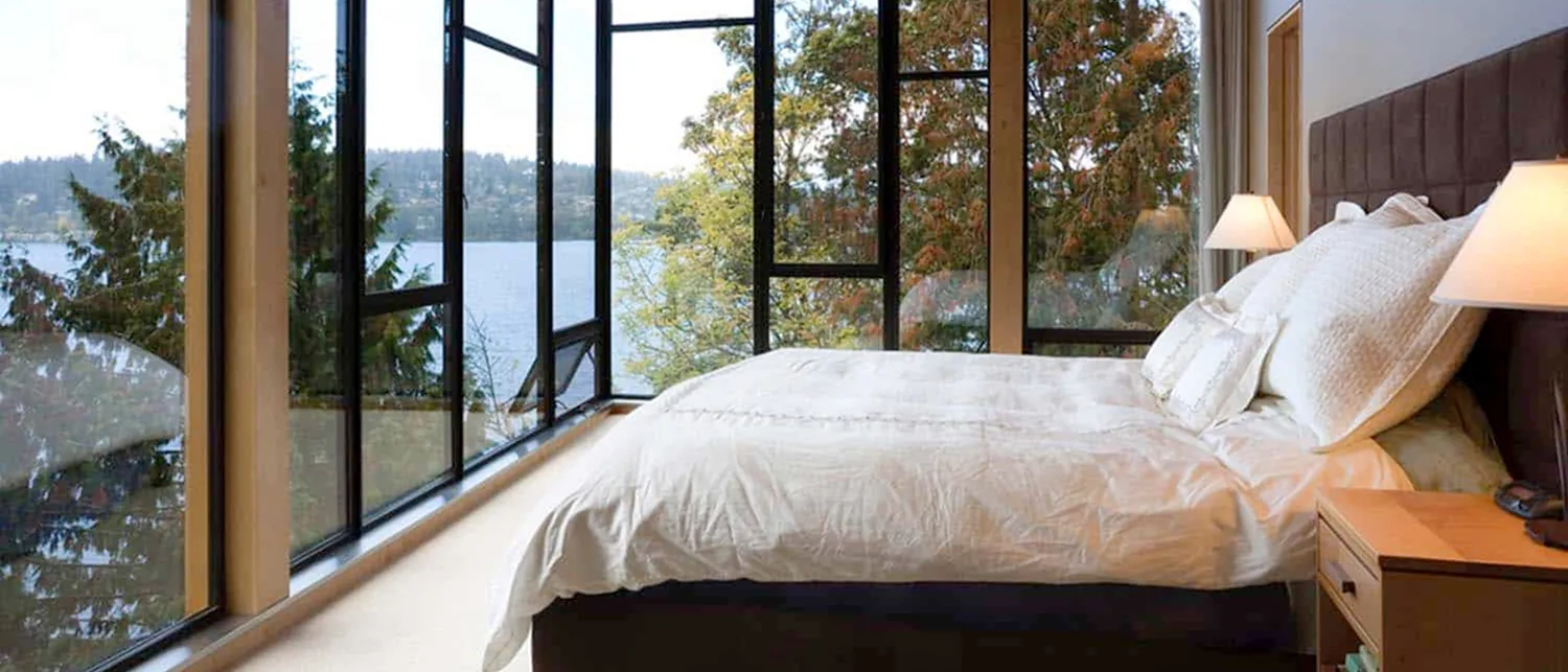 Bedroom with a wall of tall black-framed windows, one swung open, looking through autumn trees to the water.