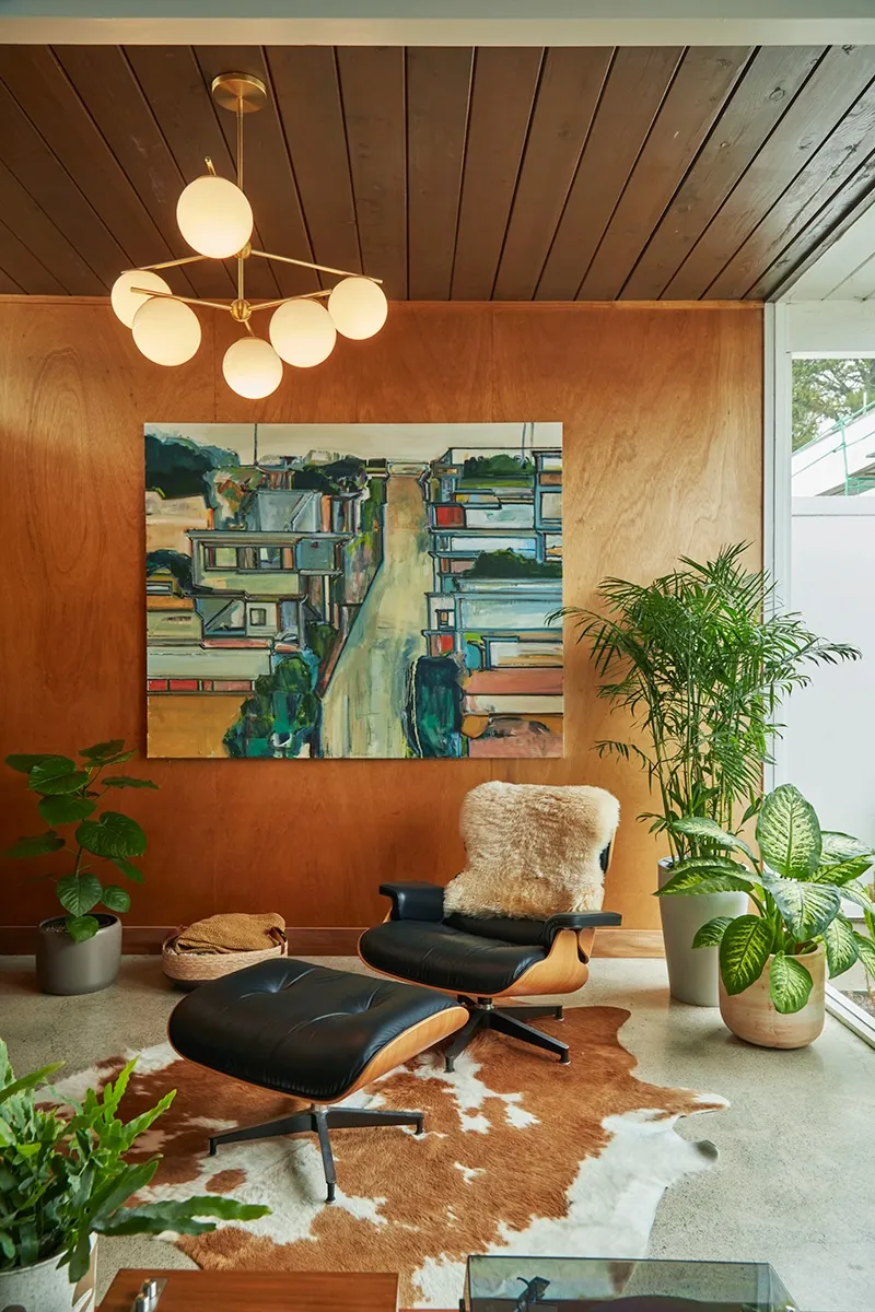 Leather lounge chair and ottoman on a cowhide rug before a plywood-panelled wall hung with a large cityscape painting.