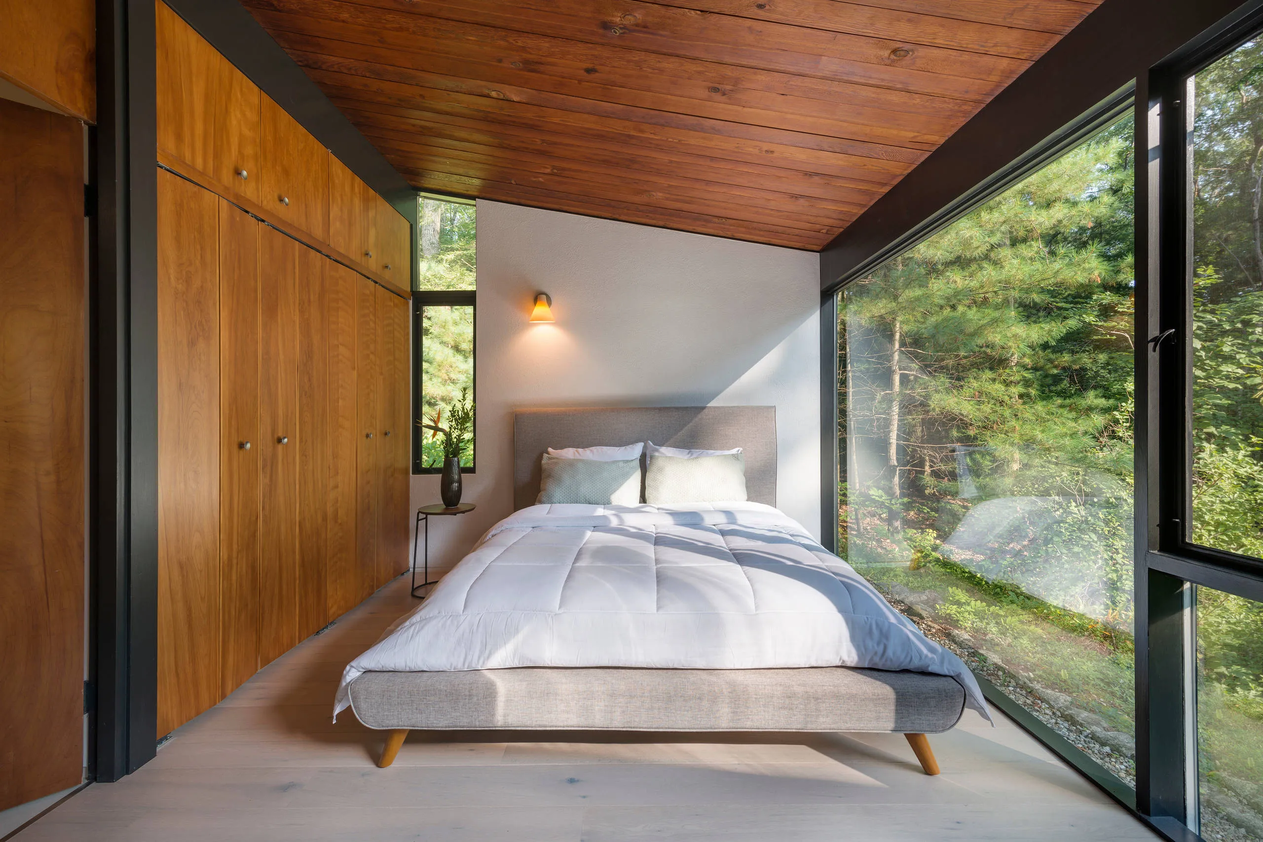 Narrow bedroom under a sloping timber ceiling, a wall of built-in wardrobes on one side and full-height glass to the woods on the other.