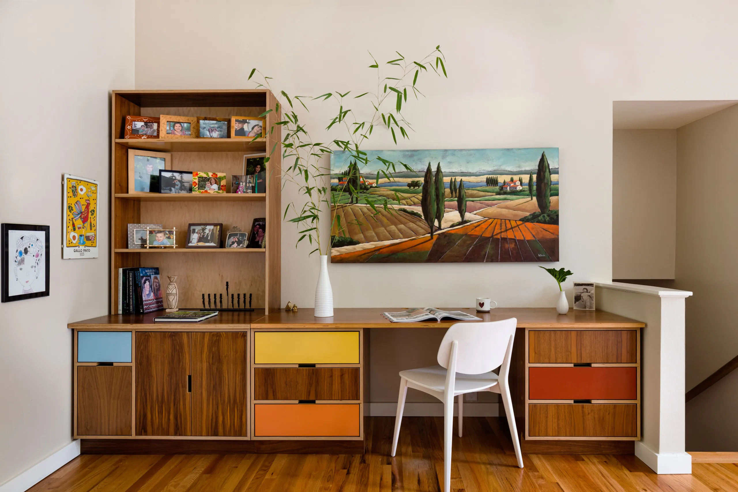 Built-in walnut desk and shelving with yellow, orange and red drawer fronts, beneath a landscape painting at the top of the stair.
