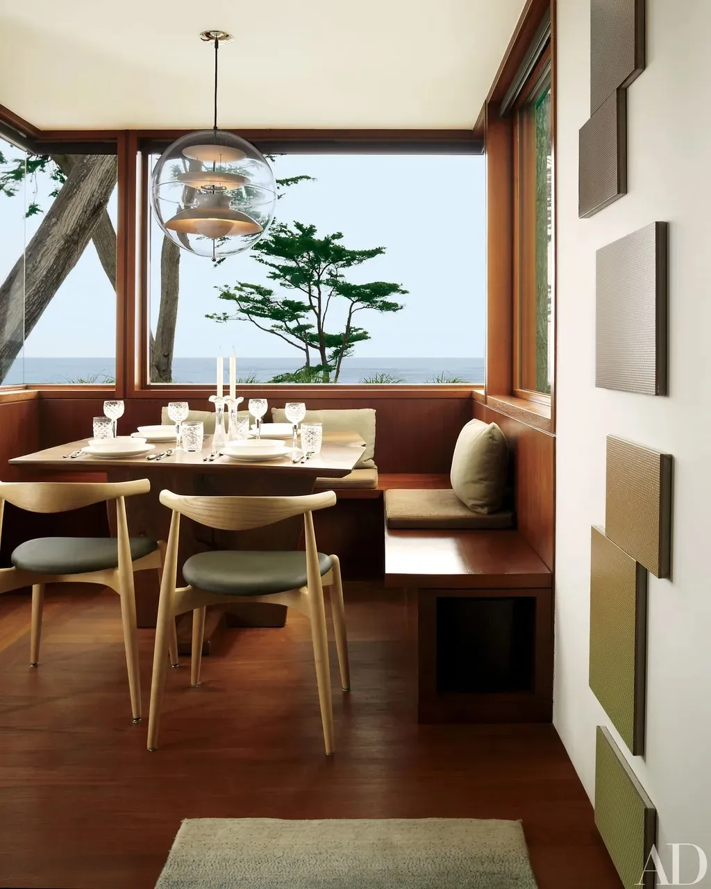 Dining nook with a built-in timber banquette and a globe pendant, its corner windows framing a windswept cypress and the sea.