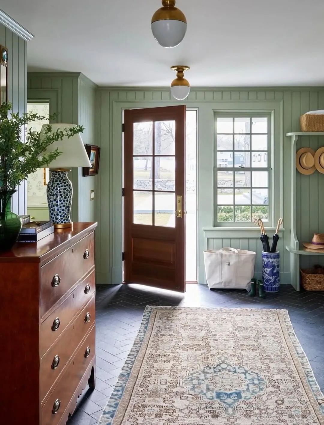 Sage-green panelled entry hall with a glazed front door standing open, a walnut chest of drawers and a patterned runner.