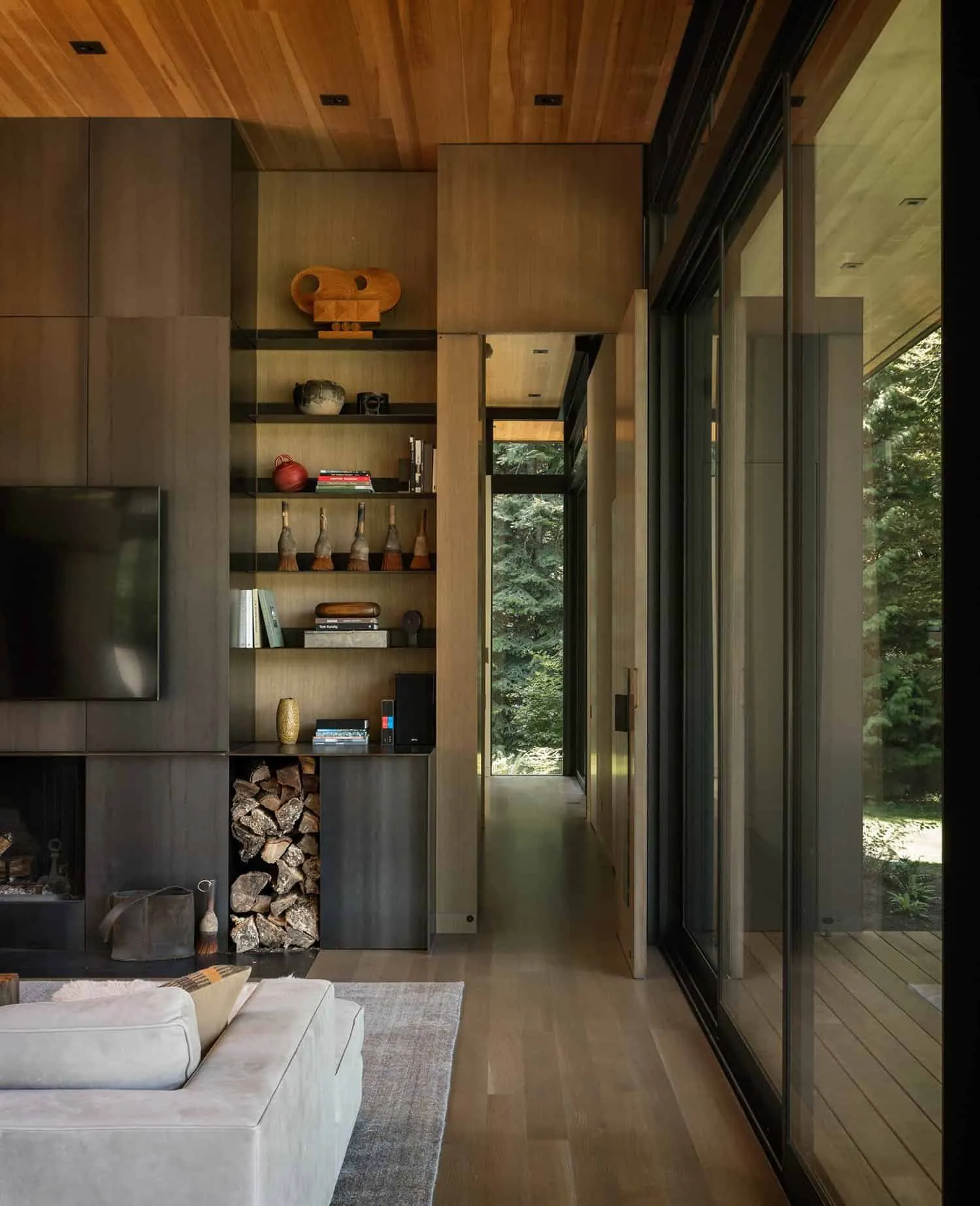 Living room with a dark timber fireplace wall, open shelving and a stacked log store beside sliding glass doors to the forest.