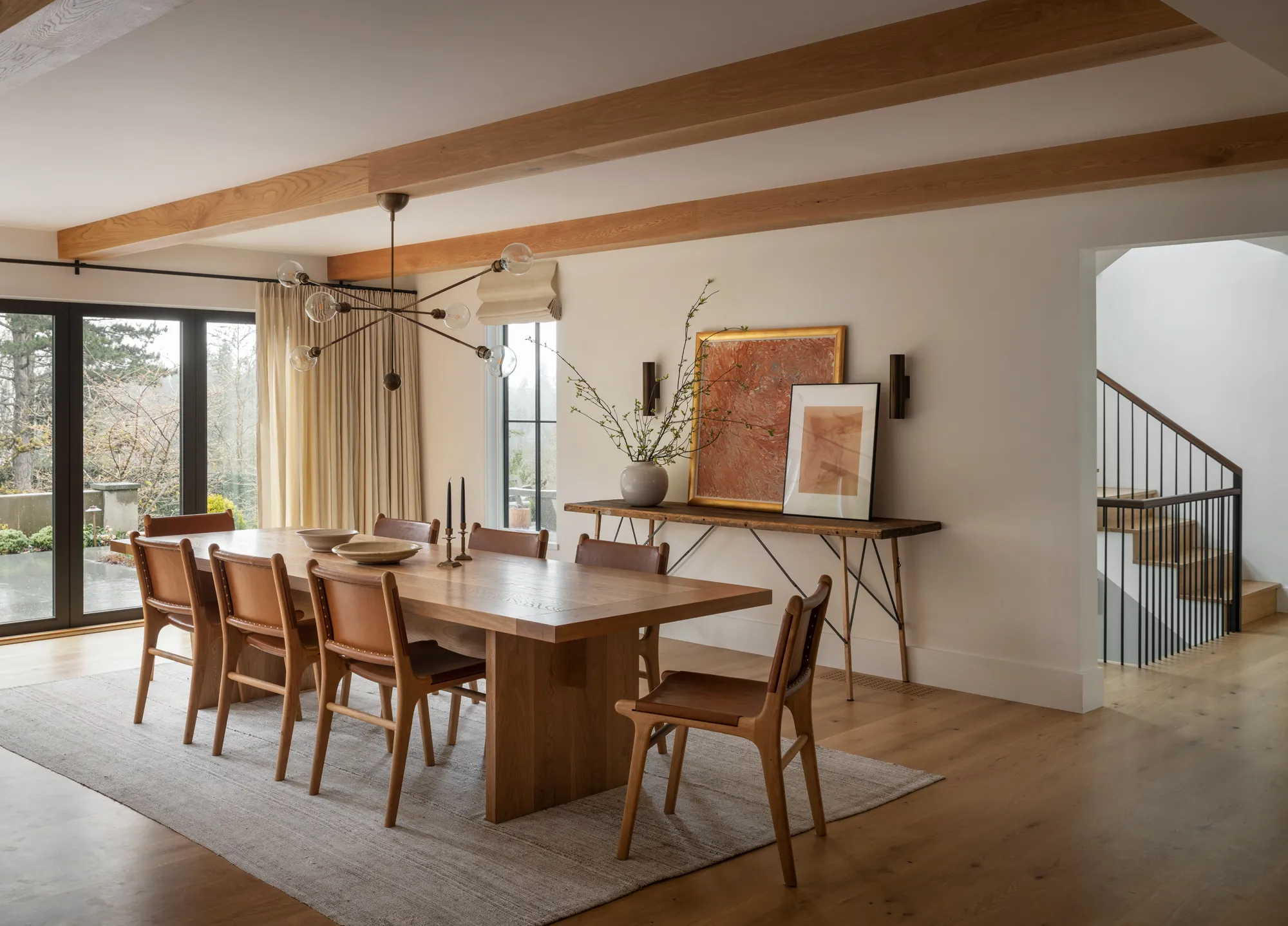 Dining room with a long oak table and leather chairs under pale timber beams, glass doors opening to a terrace.