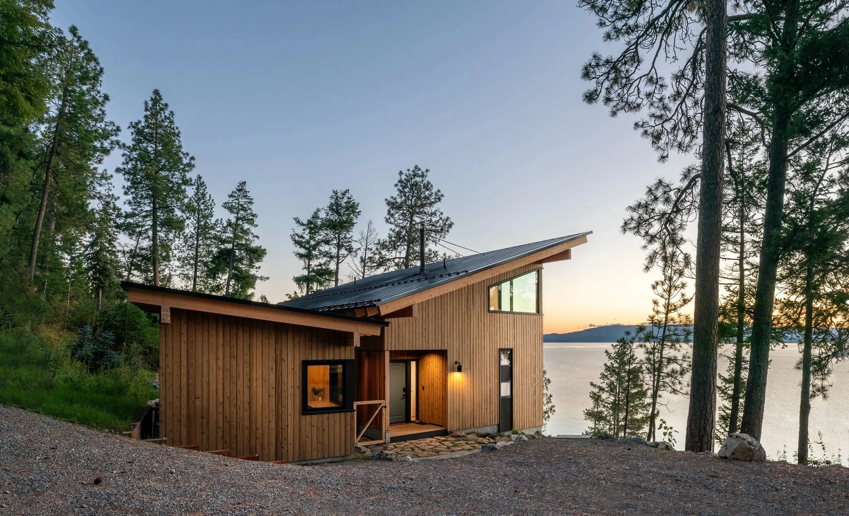 Vertical-board timber house with a steep shed roof and a high corner window, on a wooded bluff above a lake at sunset.