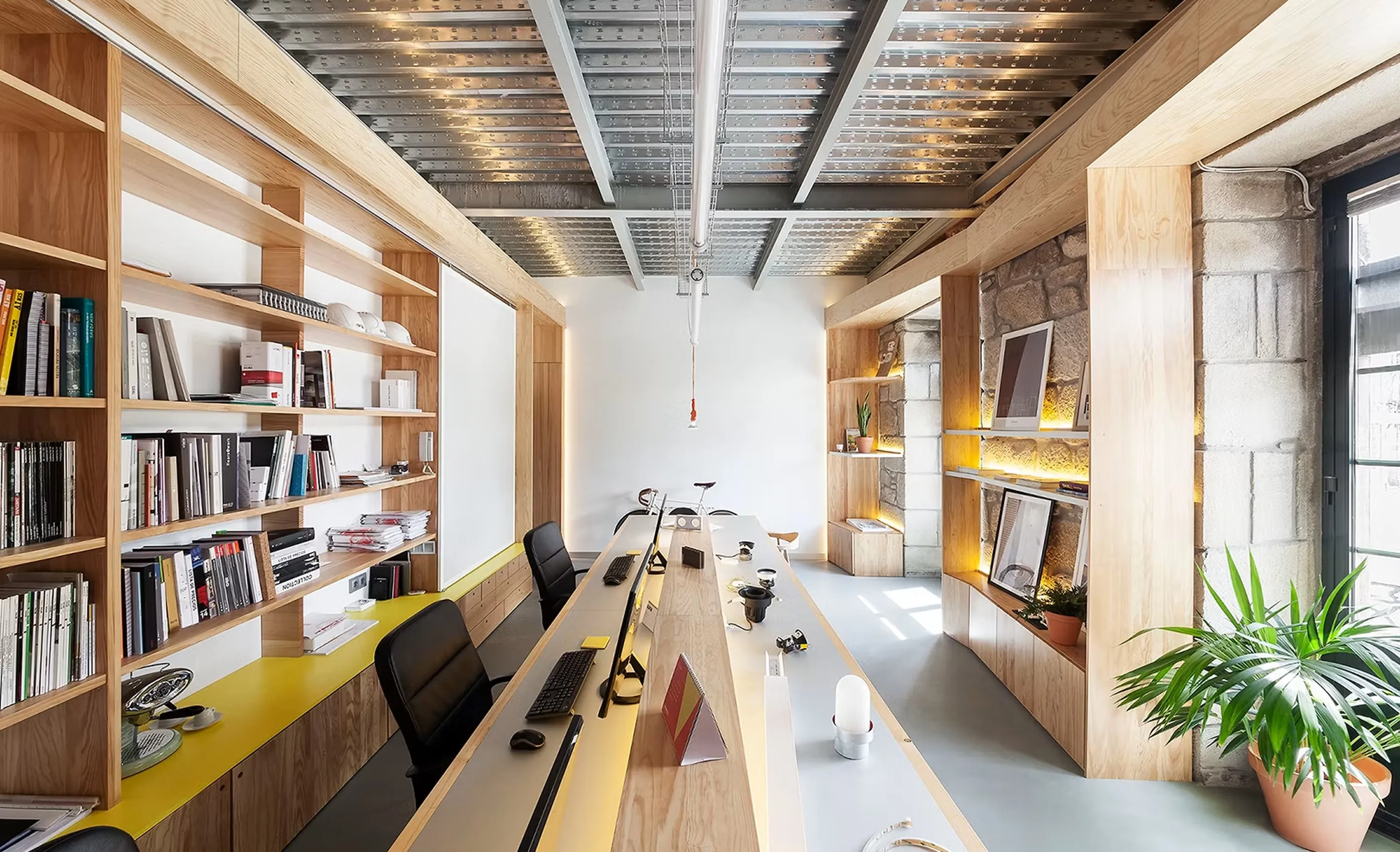 The studio: a long shared desk under an exposed steel ceiling, with timber shelving full of books and samples against a stone wall.