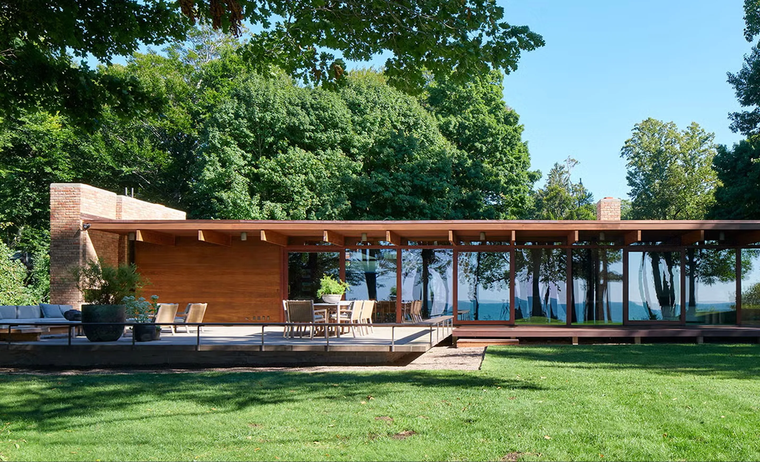 Long, low pavilion with a deep timber roof and a glass wall reflecting the trees, a brick chimney mass and a dining terrace on a lawn.