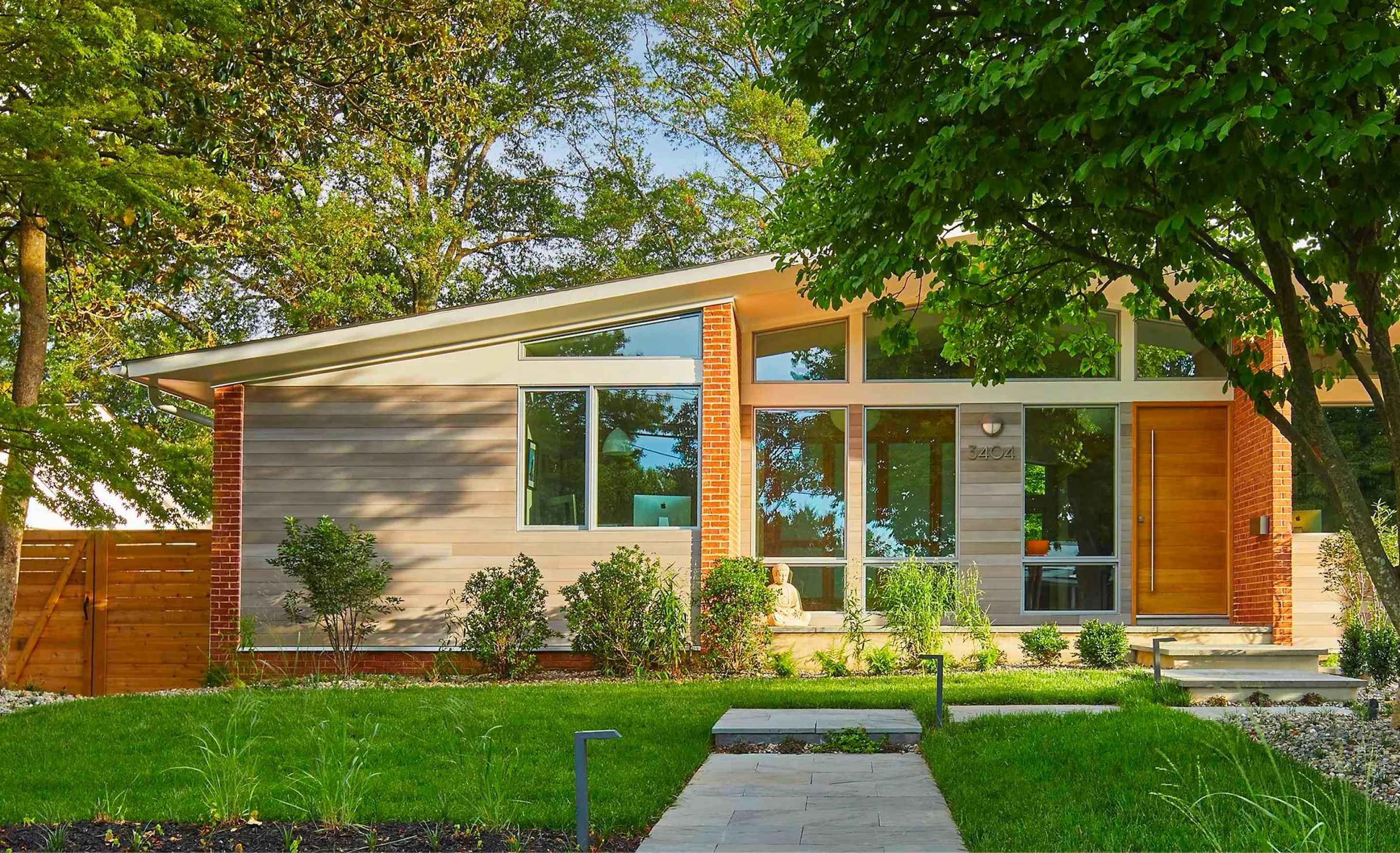Single-storey infill house with a sloping roof, brick piers, pale horizontal siding and a timber front door behind a stone path.