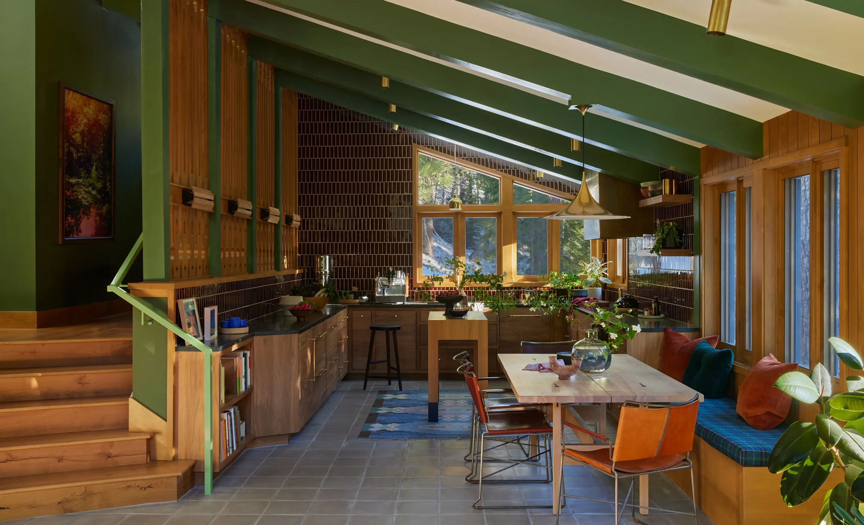 Barn kitchen and dining room under green-painted rafters, with walnut cabinets, dark tiled walls and a window seat by tall windows.