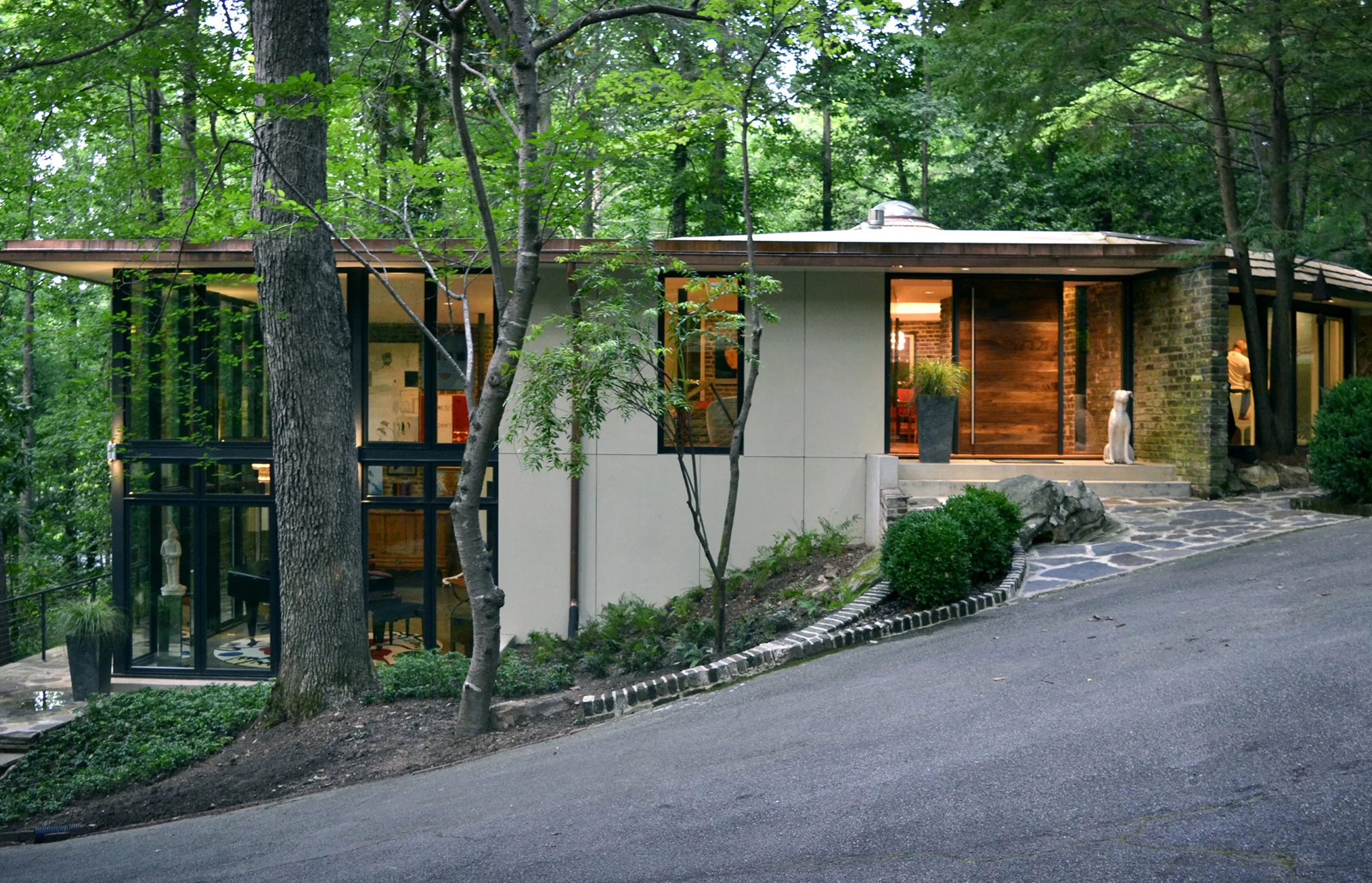 Low modern house with pale panelled walls, tall black-framed windows and a timber front door, set into a wooded slope above a curving drive.