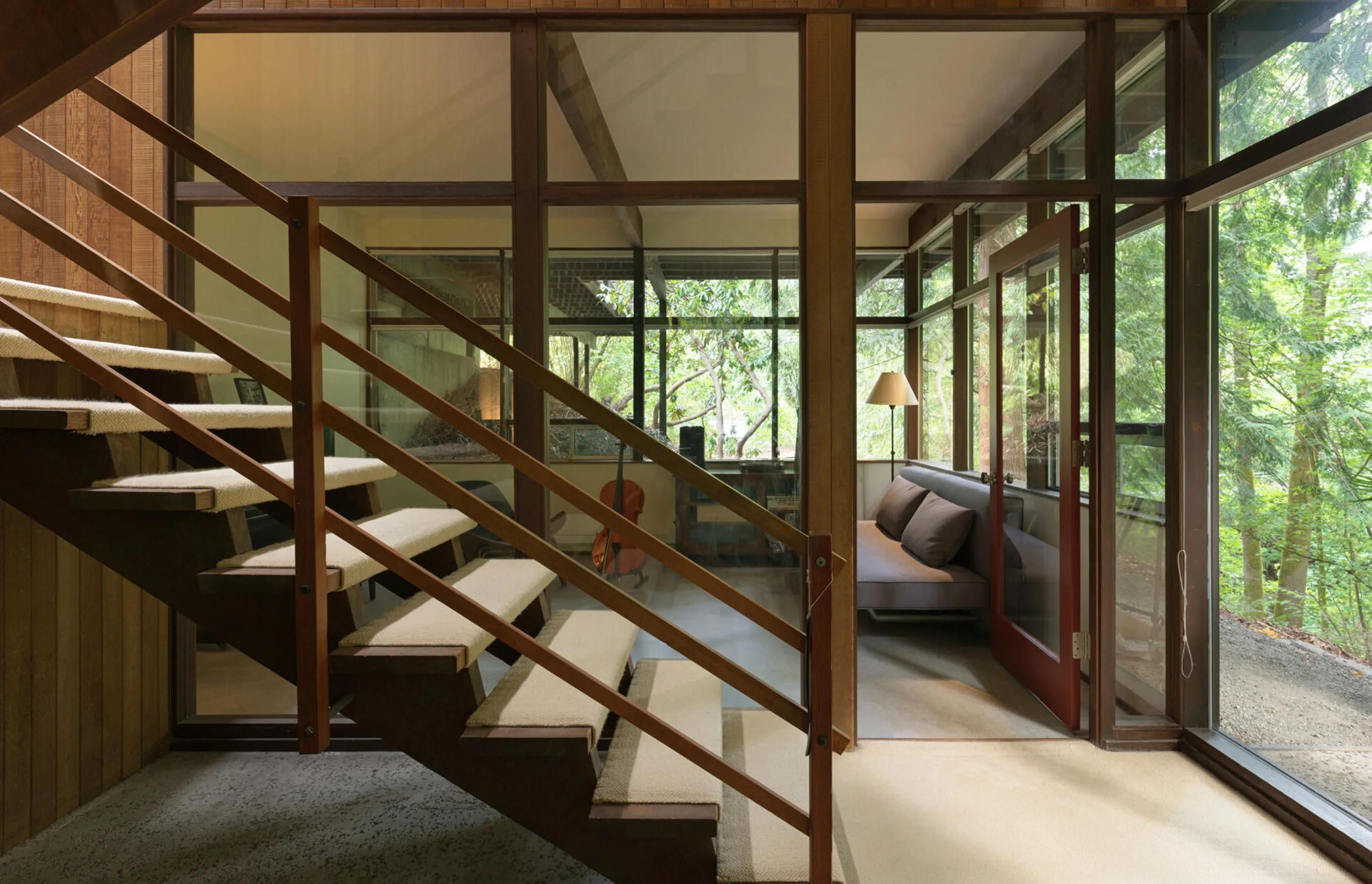 Open-tread timber stair in front of a glass-walled sitting room with a sofa and a cello, looking out into the forest.