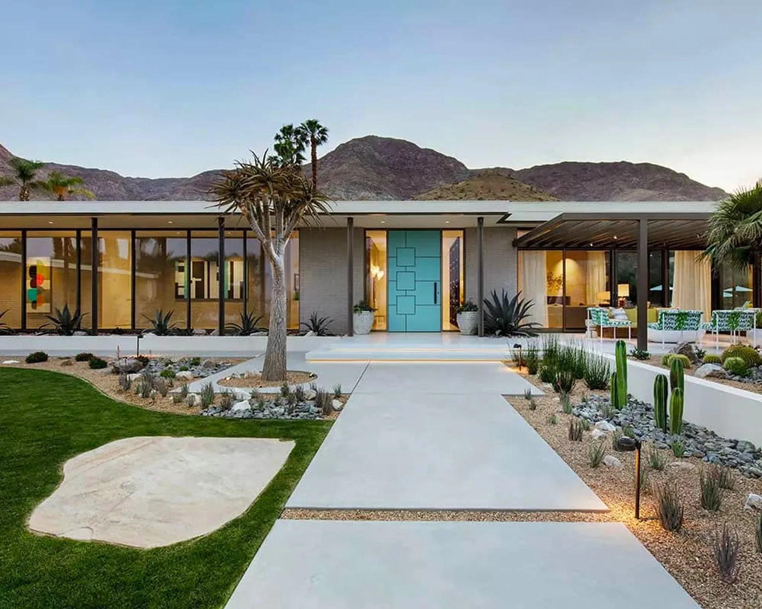 Single-storey desert house at dusk with a turquoise front door, glass walls and a concrete path, mountains behind.