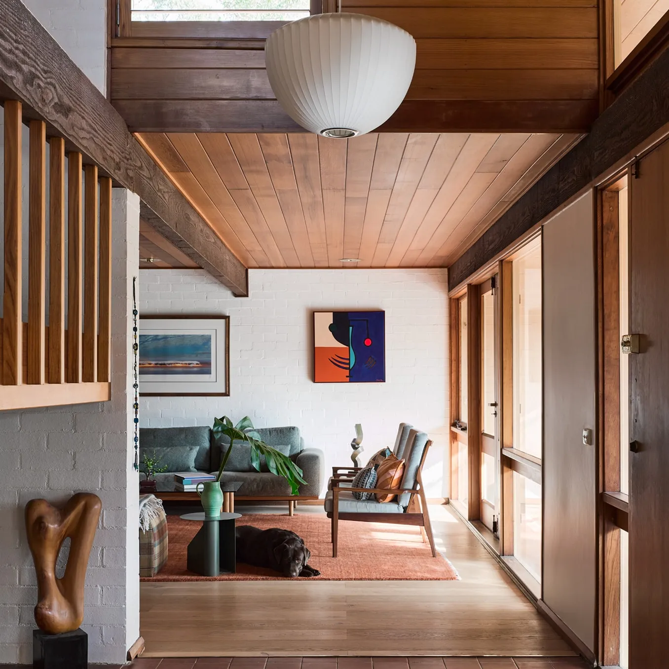 Living room under a timber-boarded ceiling, with a grey sofa and a dog asleep on a rust rug beside white-painted brick walls.