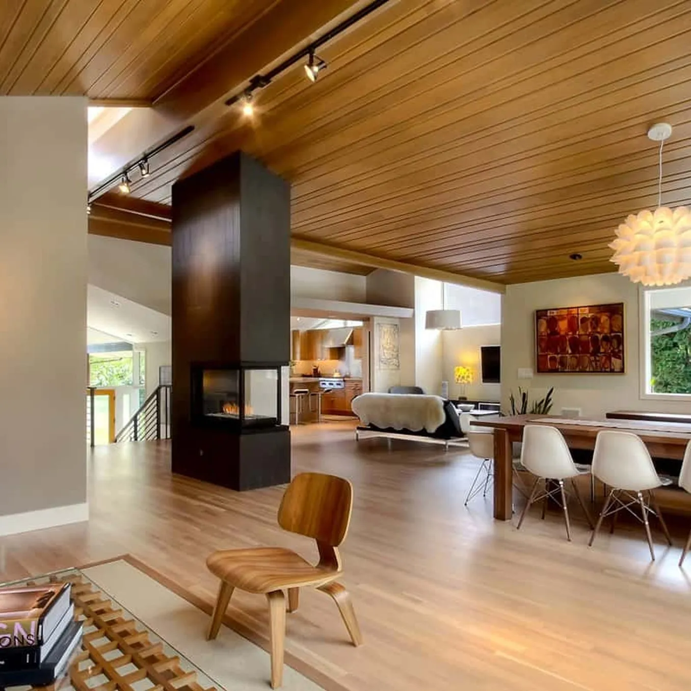 Open-plan living and dining space under a slatted timber ceiling, divided by a dark three-sided fireplace column.