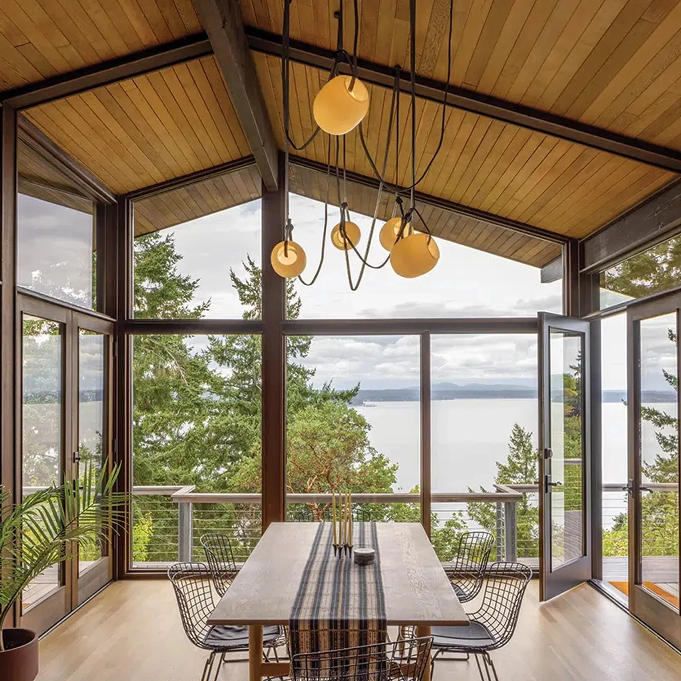 Dining room under a gabled timber ceiling, with floor-to-ceiling glass on three sides looking out over trees to open water.